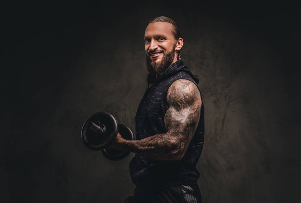 Smiling bearded tattooed athlete in a black hoodie posing with a dumbbell in hand. Isolated on dark textured background. - Photo, Image