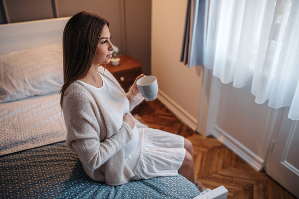 Beautiful pregnant woman sitting on the bed , holding a cup - Fotografie, Obrázek
