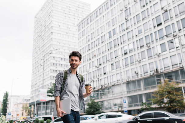 Студент-хипстер ходит в университет с ноутбуком и чашкой кофе. Позитивный бородатый мужчина в стильной повседневной одежде ходит по городской улице и позирует для фото
. - Фото, изображение