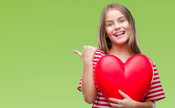 Молодая красивая девушка держит красное сердце на изолированном фоне указывая и показывая с большим пальцем вверх в сторону со счастливым лицом улыбаясь
 - Фото, изображение