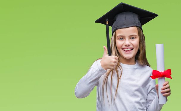Young beautiful girl wearing graduate cap holding degree over isolated background happy with big smile doing ok sign, thumb up with fingers, excellent sign - Photo, Image