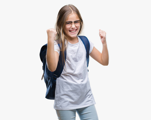 若く美しくスマートな学生女の子のバックパックを身に着けている非常に幸せと興奮の腕を上げ、勝者のジェスチャを行う分離の背景に笑みを浮かべて、成功のために叫んでいます。お祝いのコンセプト. - 写真・画像