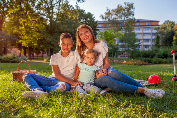Happy family spending time outdoors sittting on grass in park. Mom with two children son and daughter smiling. Family values - Photo, Image