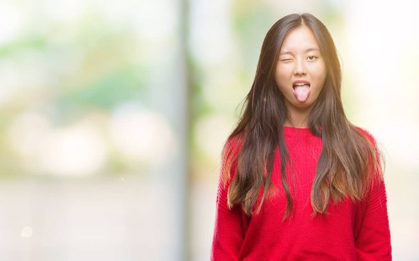 Mujer asiática joven vistiendo suéter de invierno sobre fondo aislado sacando la lengua feliz con expresión divertida. Concepto de emoción
. - Foto, imagen