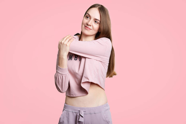 Pleased dreamy female with long dark hair, wears casual sports clothes, stretches hands, preapres for workout, stands against pink background. Active young woman trainer, being full of energy - Valokuva, kuva