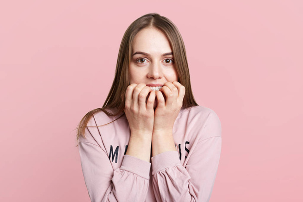 Isolated shot of pretty female model bites nails as feels very nervous and embarrassed, worries before important event, isolated over pink background. Frightened young woman looks anxiously at camera - Zdjęcie, obraz