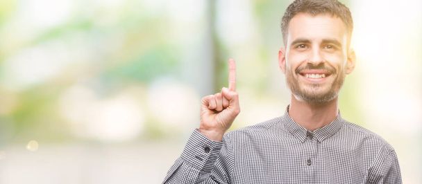 Νέοι hipster άνθρωπος δείχνει και δείχνει προς τα επάνω με ένα δάχτυλο στον αριθμό ενώ χαμογελώντας αυτοπεποίθηση και χαρούμενοι. - Φωτογραφία, εικόνα