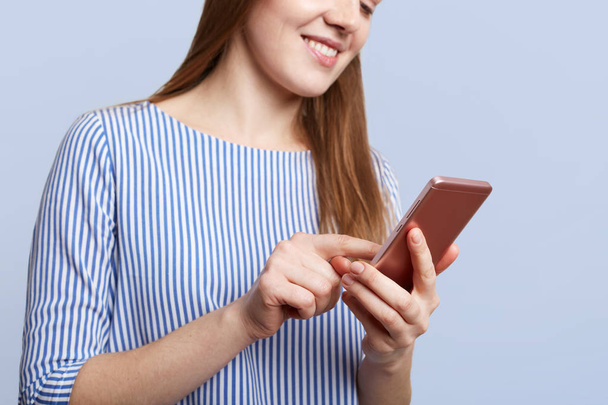 abgeschnittene Aufnahme einer jungen Frau mit fröhlichem Gesichtsausdruck hält ein modernes Handy in der Hand, Nachrichten mit Freunden, mit drahtlosem Internet verbunden, immer in Kontakt, isoliert über blauem Studiohintergrund - Foto, Bild