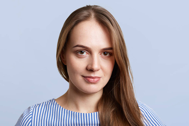 Close up portrait of brunette female with pleasant look, looks directly into camera with serious expression, isolated over light blue background. People, beauty, youth and facial expressions concept - Foto, imagen