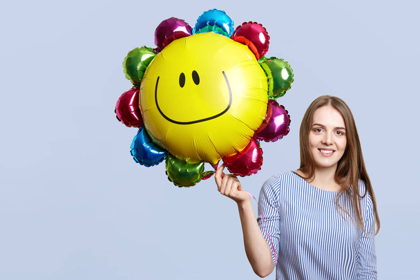 Glad brunette woman wears striped blouse, holds balloon, happy to celebrate daughter`s birthday, meets guests, isolated over blue background. Festive mood. Joyful female with air balloon sun - Foto, imagen