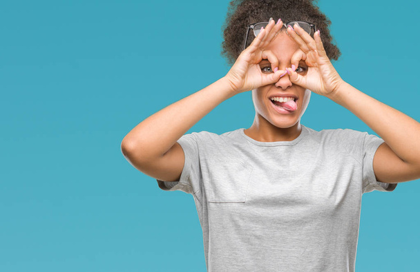 Mujer afroamericana joven con gafas sobre un fondo aislado haciendo un buen gesto como prismáticos sacando la lengua, ojos mirando a través de los dedos. Expresión loca
. - Foto, imagen