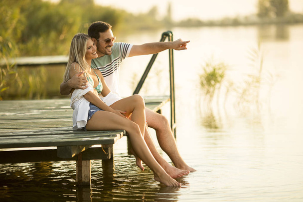 Ρομαντικό ζευγάρι κάθεται στην ξύλινη προβλήτα στη λίμνη σε ηλιόλουστη ημέρα - Φωτογραφία, εικόνα