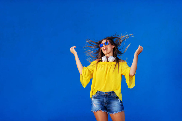 Cheerful girl in yellow shirt standing against blue wall with headset around her neck. Dancing and smiling. - Photo, Image
