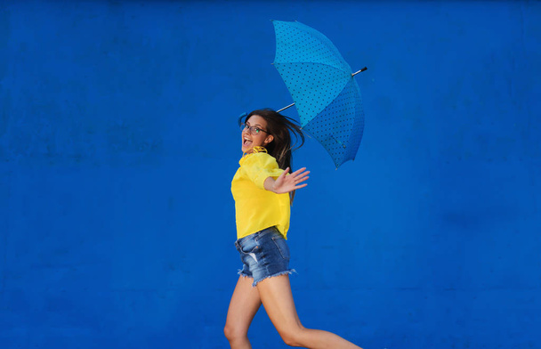 Vrolijke jonge meisje in gele overhemd blauwe paraplu houden en permanent tegen blauwe muur. - Foto, afbeelding
