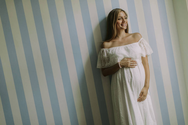 Kaunis nuori raskaana oleva nainen seisoo seinän vieressä huoneessa
 - Valokuva, kuva