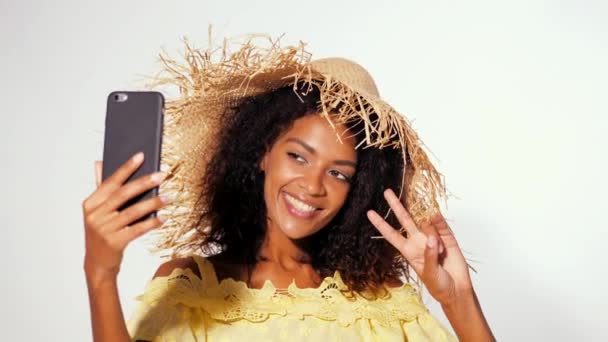 Sorrindo mulher americana africana feliz com cabelo encaracolado vestido amarelo e chapéu de palha fazendo selfie no smartphone sobre fundo da parede branca. Menina se divertindo, constrói rostos, brincando
. - Filmagem, Vídeo