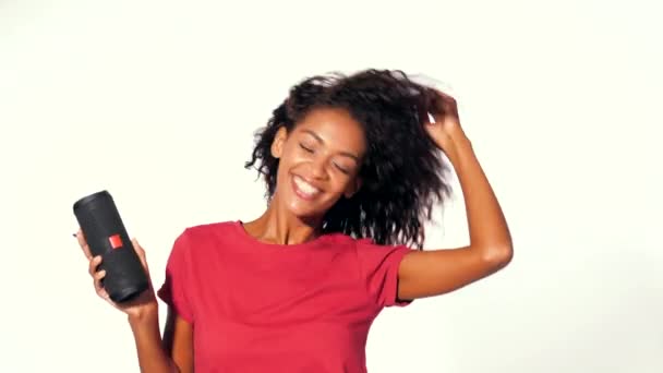 Young beautiful african american woman enjoying and dancing at white background. Modern trendy girl with afro hairstyle listening to music by wireless portable speaker. - Filmagem, Vídeo