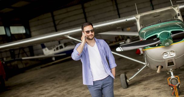 Young handsome young pilot checking airplane in the hangar and using mobile phone - Foto, immagini