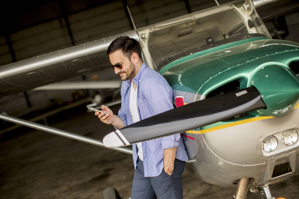 Молодой красивый молодой пилот проверяет самолет в ангаре и пользуется мобильным телефоном
 - Фото, изображение