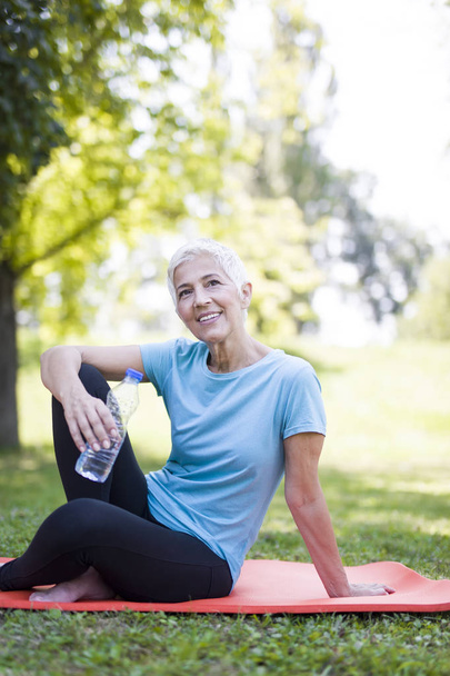 Portrait of smiling senior woman relaxing after exercising while sitting on grass - Photo, Image