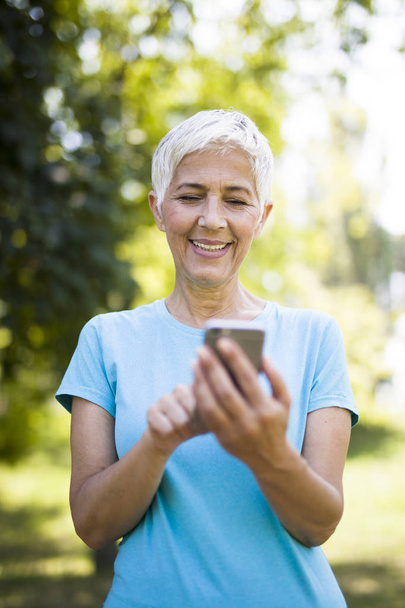 Portrét sportovní starší ženy pomocí mobilního telefonu v parku - Fotografie, Obrázek