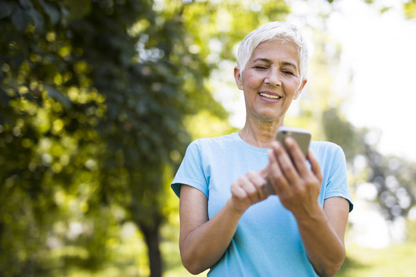 Портрет спортивной пожилой женщины с помощью мобильного телефона в парке
 - Фото, изображение