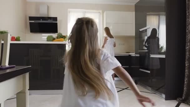 Small girl happy meeting her older sister, who cut vegetables for prepare vegan food in the kitchen at home. Child running into sisters hands to hug her - Video