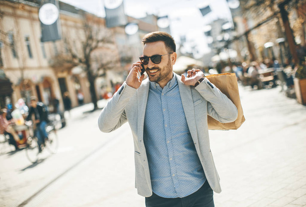 Πορτρέτο του χαρούμενα όμορφος άνδρας πηγαίνει ψώνια στην πόλη ενώ χρησιμοποιεί κινητό τηλέφωνο - Φωτογραφία, εικόνα