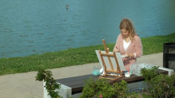 A pretty woman with red hair, paints a picture on canvas, which stands on the easel. The lady is in the open air near the lake of the river, she draws from life - Footage, Video