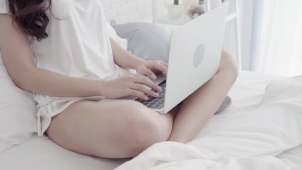 Slow motion - Beautiful Asian mix race Caucasian woman using computer or laptop while lying on bed in her bedroom. Happy female buying online shopping at home. Lifestyle woman relaxing at home concept - Πλάνα, βίντεο