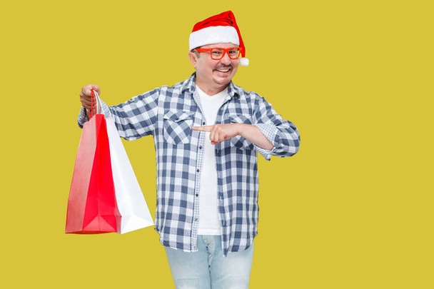 Happy middle aged man in red santa cap holding shopping packages on yellow background - Foto, Bild