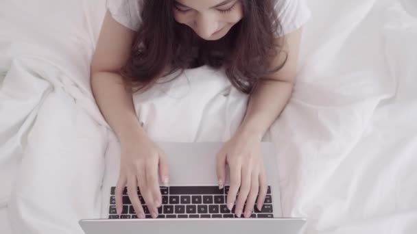 Beautiful Asian mix race Caucasian woman using computer or laptop while lying on the bed in her bedroom. Happy female buying online shopping at home. Lifestyle woman relaxing at home concept. - Video