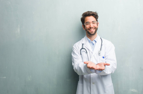 Young friendly doctor man against a grunge wall with a copy space holding something with hands, showing a product, smiling and cheerful, offering an imaginary object - Photo, image
