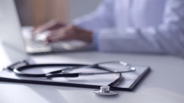 gedeeltelijke weergave van arts werkzaam met laptop met de stethoscoop en Klembord liggend op de voorgrond - Video
