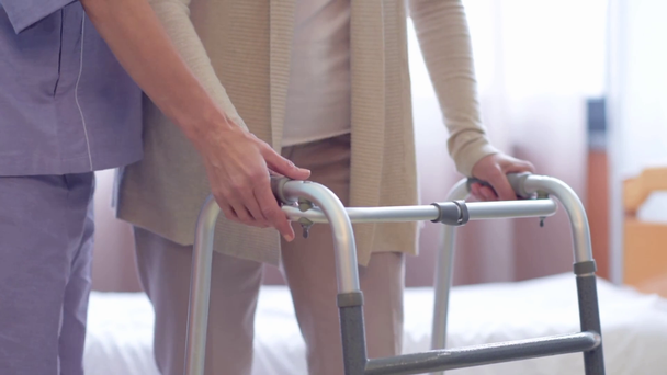 частковий вид медсестри, що підтримує старшу жінку, коли вона встає з ліжка і бере свою прогулянкову рамку
 - Кадри, відео