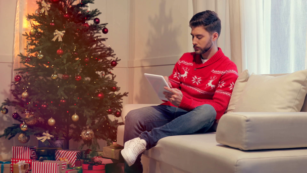 joven guapo sentado en el sofá y el uso de la tableta en la sala de estar decorada de Navidad
 - Metraje, vídeo