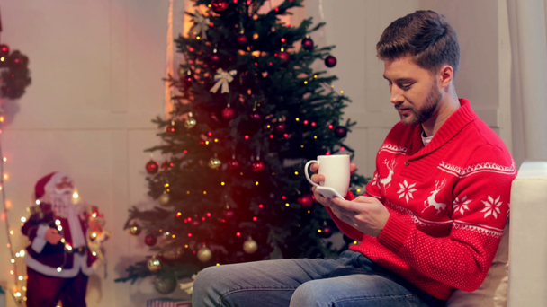 boldog, fiatal ember ül a kanapén csésze kávé és használ smartphone, karácsonyi díszített nappali - Felvétel, videó