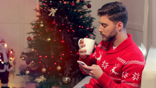 joven guapo sentado en el sofá con una taza de café y el uso de teléfono inteligente en la sala de estar decorada con Navidad
 - Metraje, vídeo