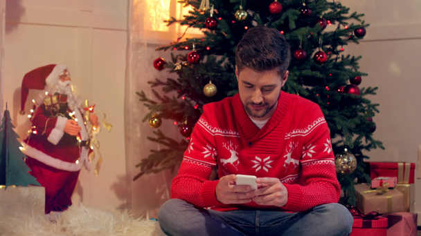 handsome young man sitting on floor and using smartphone in christmas decorated living room - Séquence, vidéo