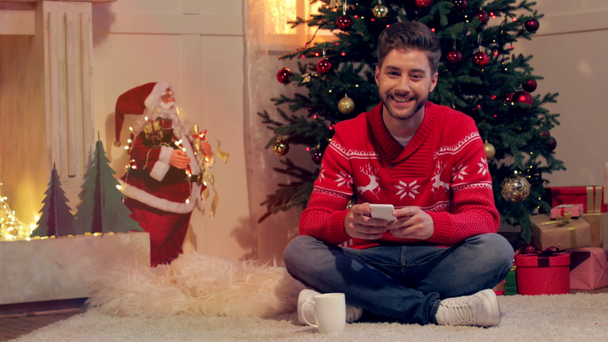 atractivo joven sentado en el suelo y el uso de teléfono inteligente en la sala de estar decorada con navidad
 - Metraje, vídeo