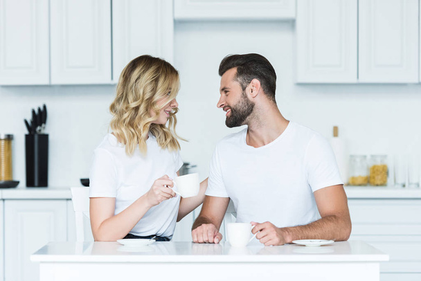 happy young couple drinking coffee and smiling each other in kitchen at morning - Fotografie, Obrázek