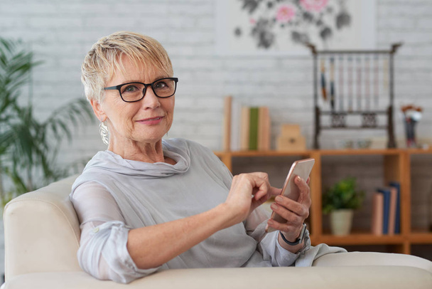 Elderly woman sitting on sofa and using application in her smartphone - Photo, Image