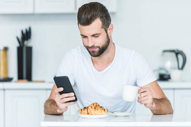 handsome bearded young man using smartphone during breakfast - Photo, Image