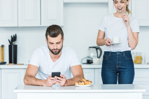 young man using smartphone while girlfriend holding cup of coffee behind - Foto, Imagem