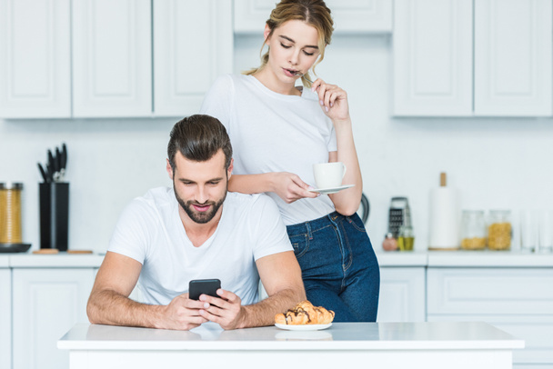 attractive young woman holding cup of coffee and looking at boyfriend using smartphone - Foto, immagini