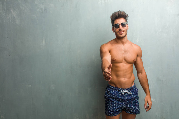 junger, athletischer Mann im Badeanzug vor einer Grunge-Wand, der sich ausstreckt, um jemanden zu grüßen oder um zu helfen gestikuliert, glücklich und aufgeregt - Foto, Bild
