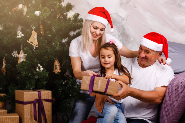 iäkäs mies Santa hattu viettää aikaa tyttärentytär ja tytär lähellä joulukuusi
 - Valokuva, kuva