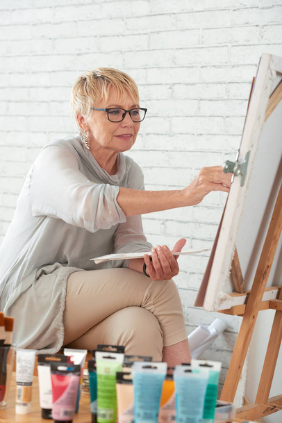 Старая креативная женщина в очках, сосредоточенная на живописи
 - Фото, изображение