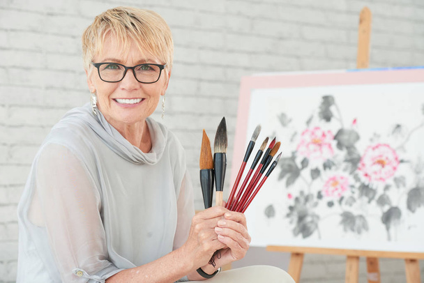 Portrait of happy aged woman showing her brushes for painting - Foto, Imagem
