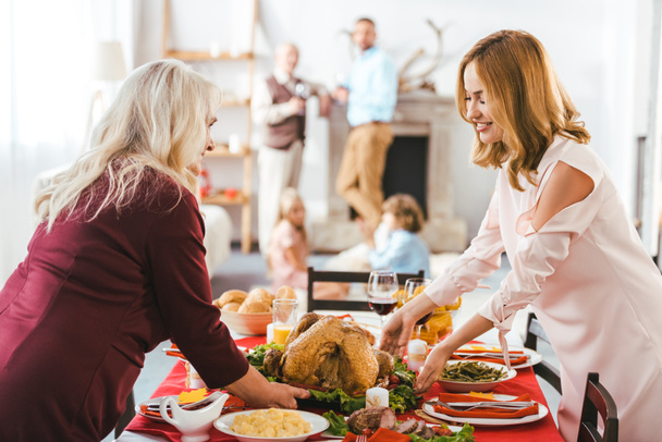 smiling young and senior women serving thanksgiving day table while men and kids standing blurred on background - Φωτογραφία, εικόνα